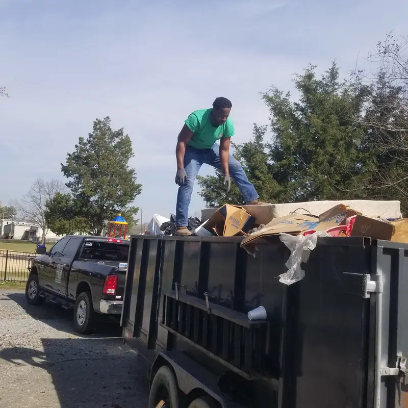 A crew member in a green Racoons shirt standing on a loaded dump trailer beside the pickup