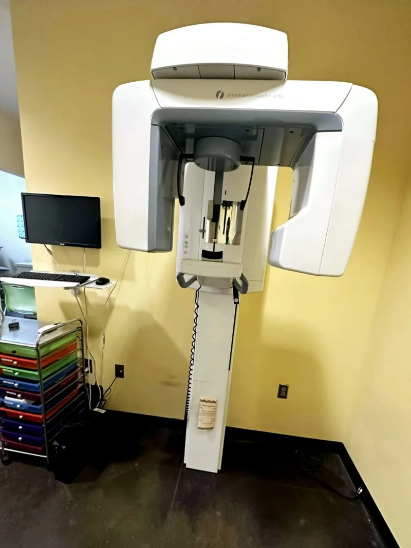 A dental panoramic X-ray unit still mounted in the treatment room next to its monitor and supply cart