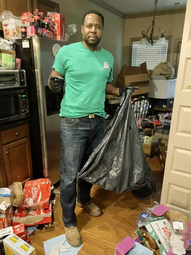 A crew member hauling a bag of trash out of a cluttered kitchen
